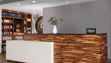 A hotel reception desk with a modern design, a sign indicating it's a Hilton Honors location, and a variety of snacks and beverages on shelves.