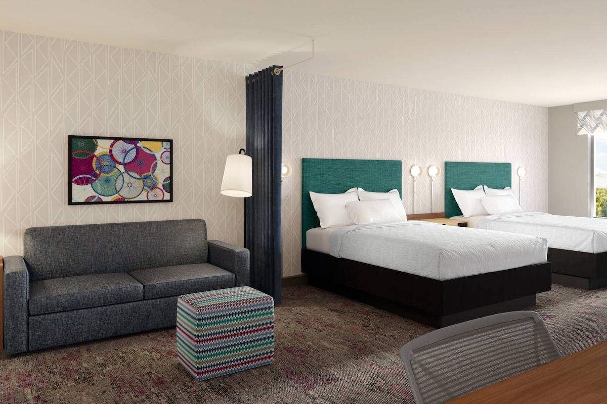A hotel room with a gray sofa, two beds with green headboards, a colorful abstract painting, and a patterned carpet.