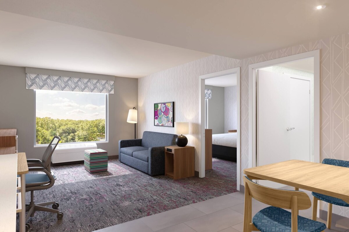 A hotel room with a desk, a chair, a sofa, a small table, a lamp, a rug, and a painting on the wall.