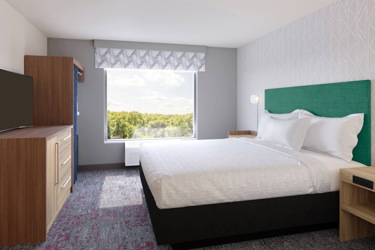 A modern hotel room with a large bed, a television, a desk, and a window with a view of trees.