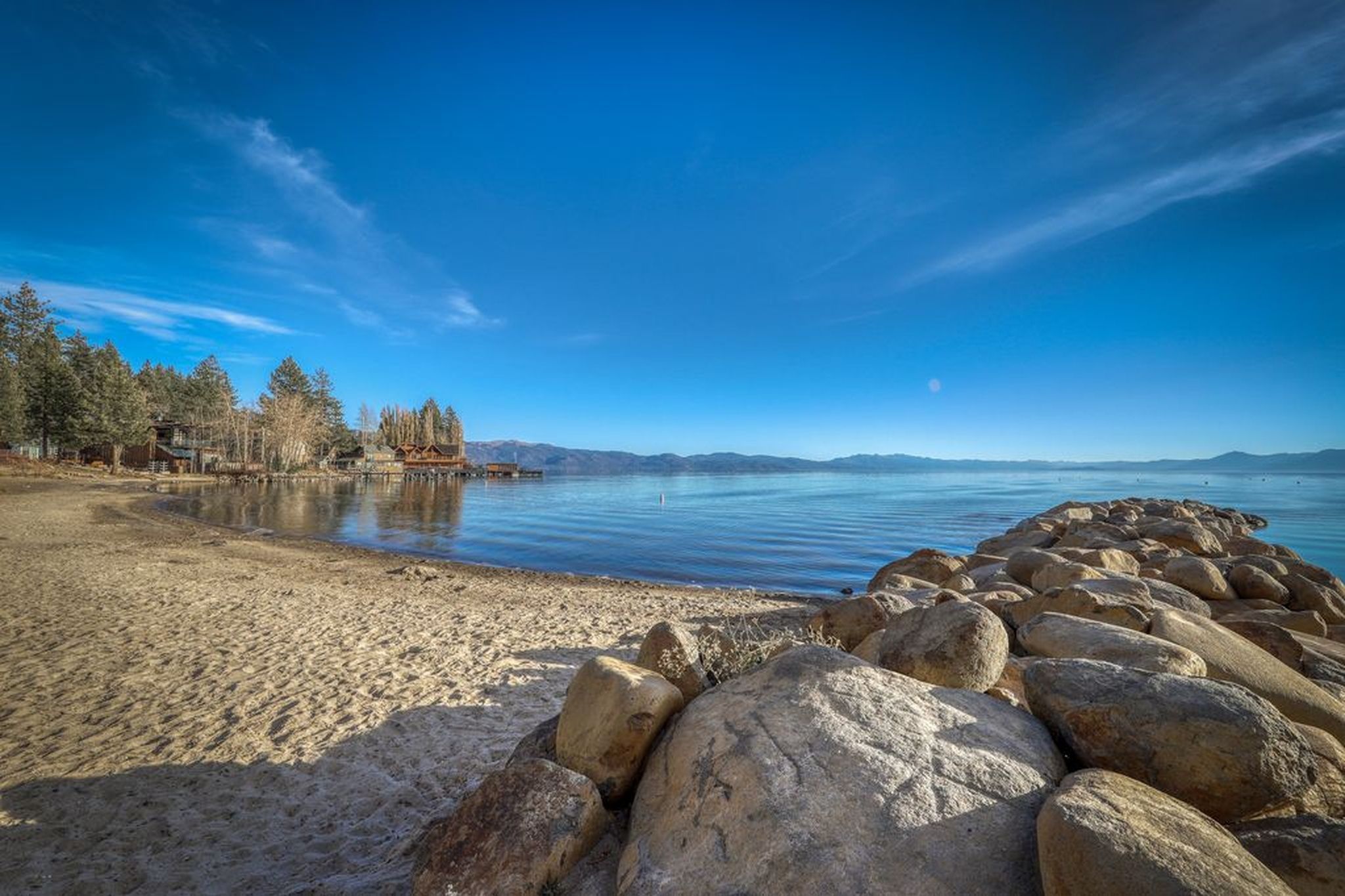 A beach with a rocky shoreline, calm water, and a forested area with buildings in the background.