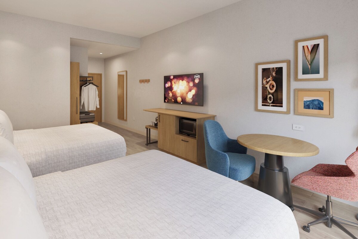 Una habitación de hotel con dos camas, un escritorio, una silla, un televisor y obras de arte en la pared.