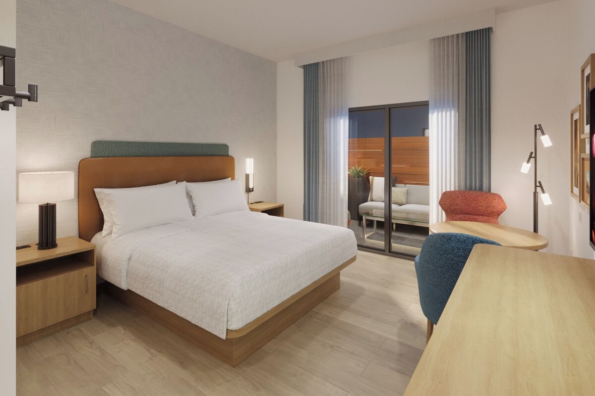 Una habitación de hotel con cama, mesitas de noche, un escritorio, una silla y un amplio ventanal con cortinas.
