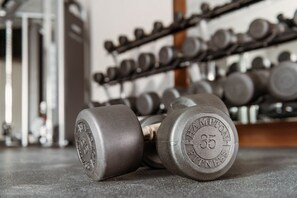Un par de mancuernas con inscripciones sobre ellas, colocadas en el suelo de un gimnasio con un estante de pesos similares al fondo.