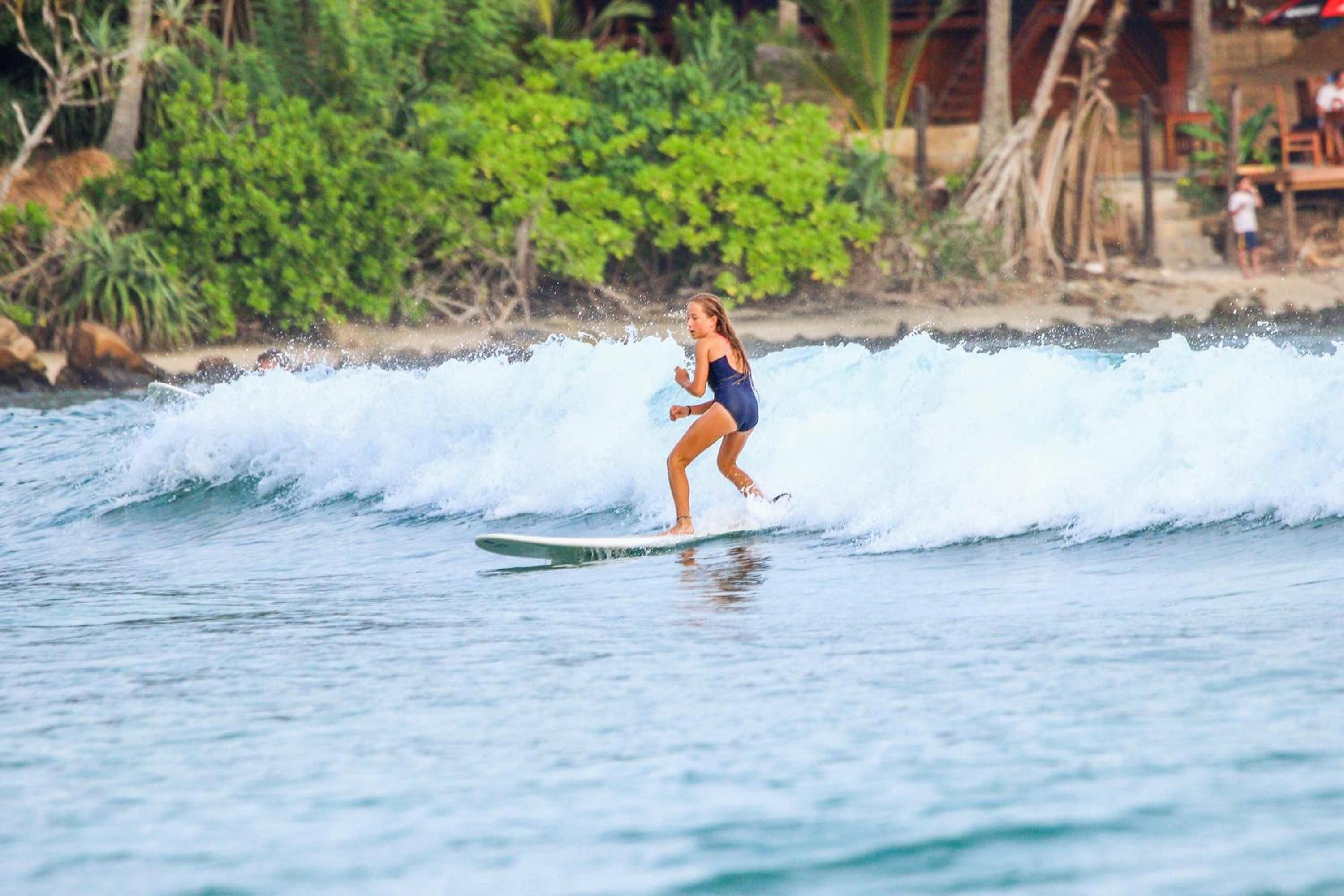 A person surfing on a wave with a beach and greenery in the background.