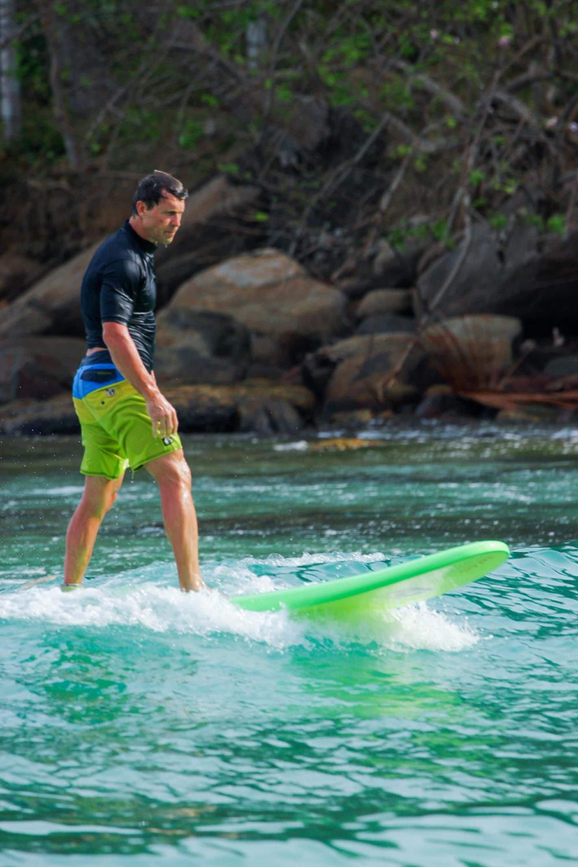 A person standing on a surfboard in a body of water with rocks and greenery in the background.
