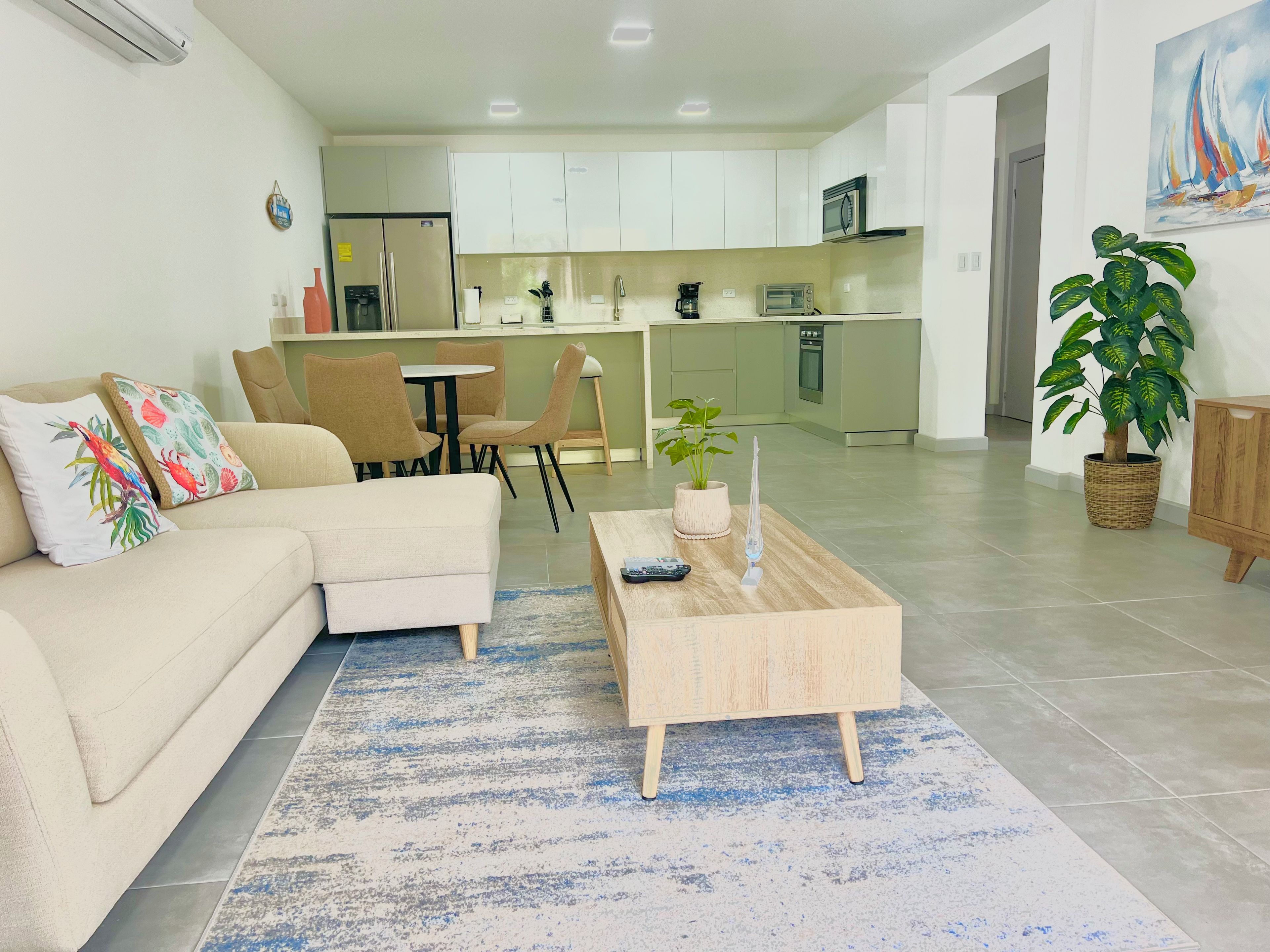 A modern living room with a sofa, coffee table, and chairs. A kitchen is visible in the background with white cabinets and appliances.