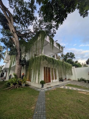 A two-story house with a covered porch, a paved pathway, and a large tree.