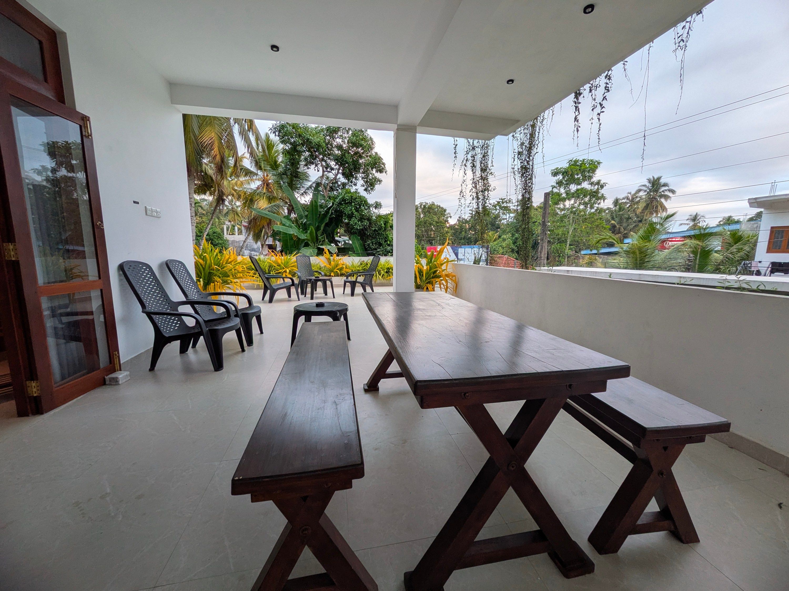 Un patio al aire libre con mesas y sillas de madera, rodeado de vegetación y con vistas a una zona residencial.
