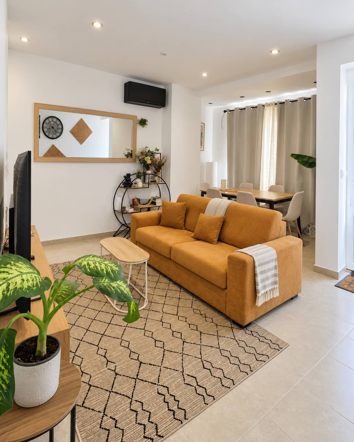 Una sala de estar moderna con sofá, una mesa de centro y televisor. Hay un comedor con mesa y sillas. La habitación tiene una alfombra con estampado geométrico y una planta grande.