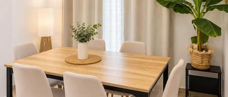 A dining area with a wooden table, four chairs, a vase with greenery, a lamp, and a large potted plant.