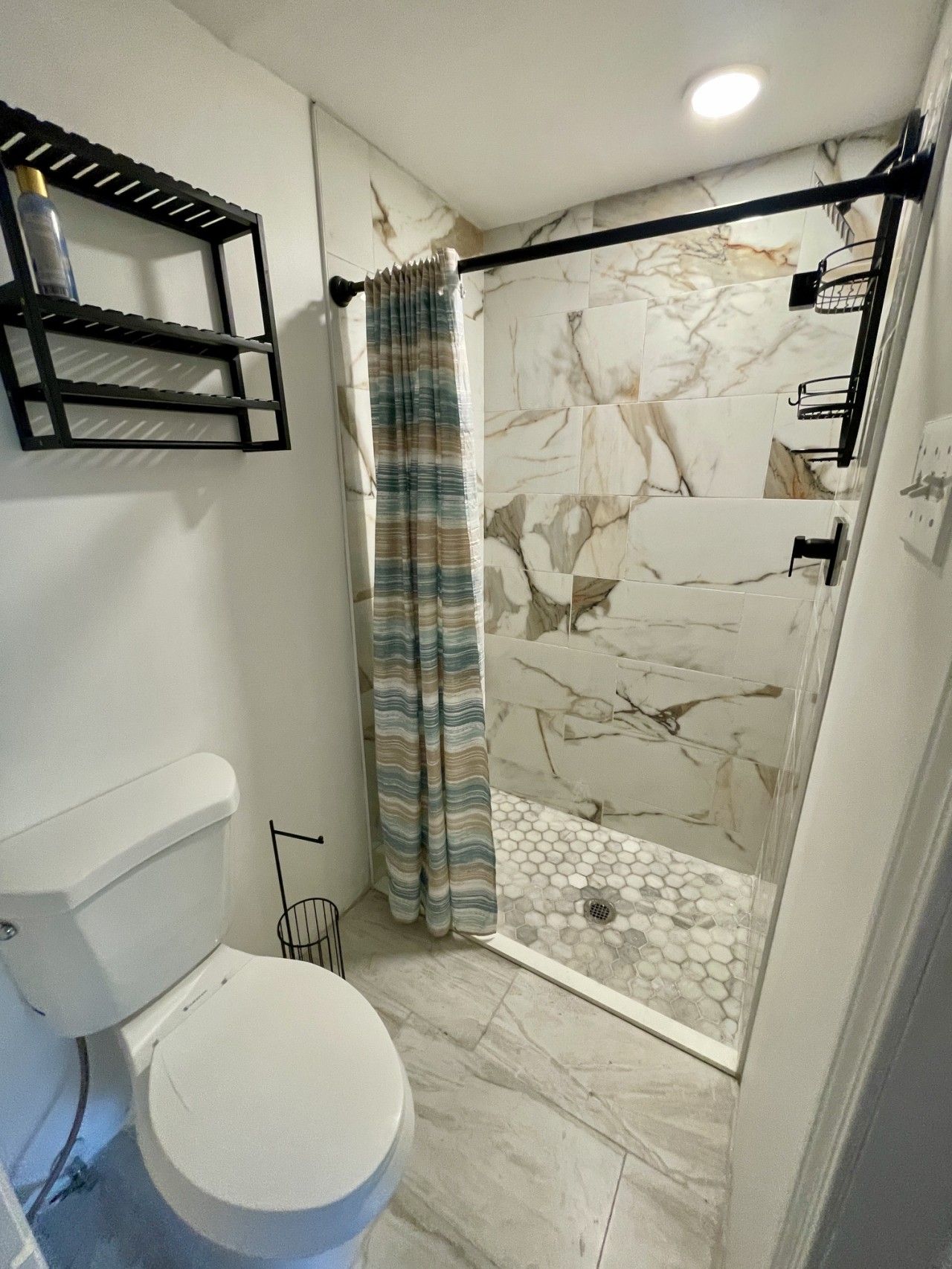 A bathroom with a toilet, a shower with a marble wall, and a shelf unit.