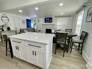 A modern kitchen with a central island, dining area with chairs, and a television mounted above the fireplace.