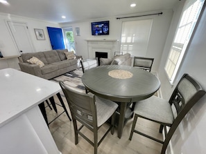 A modern living room with a grey sofa, a round dining table, and a flat-screen TV mounted above a fireplace.