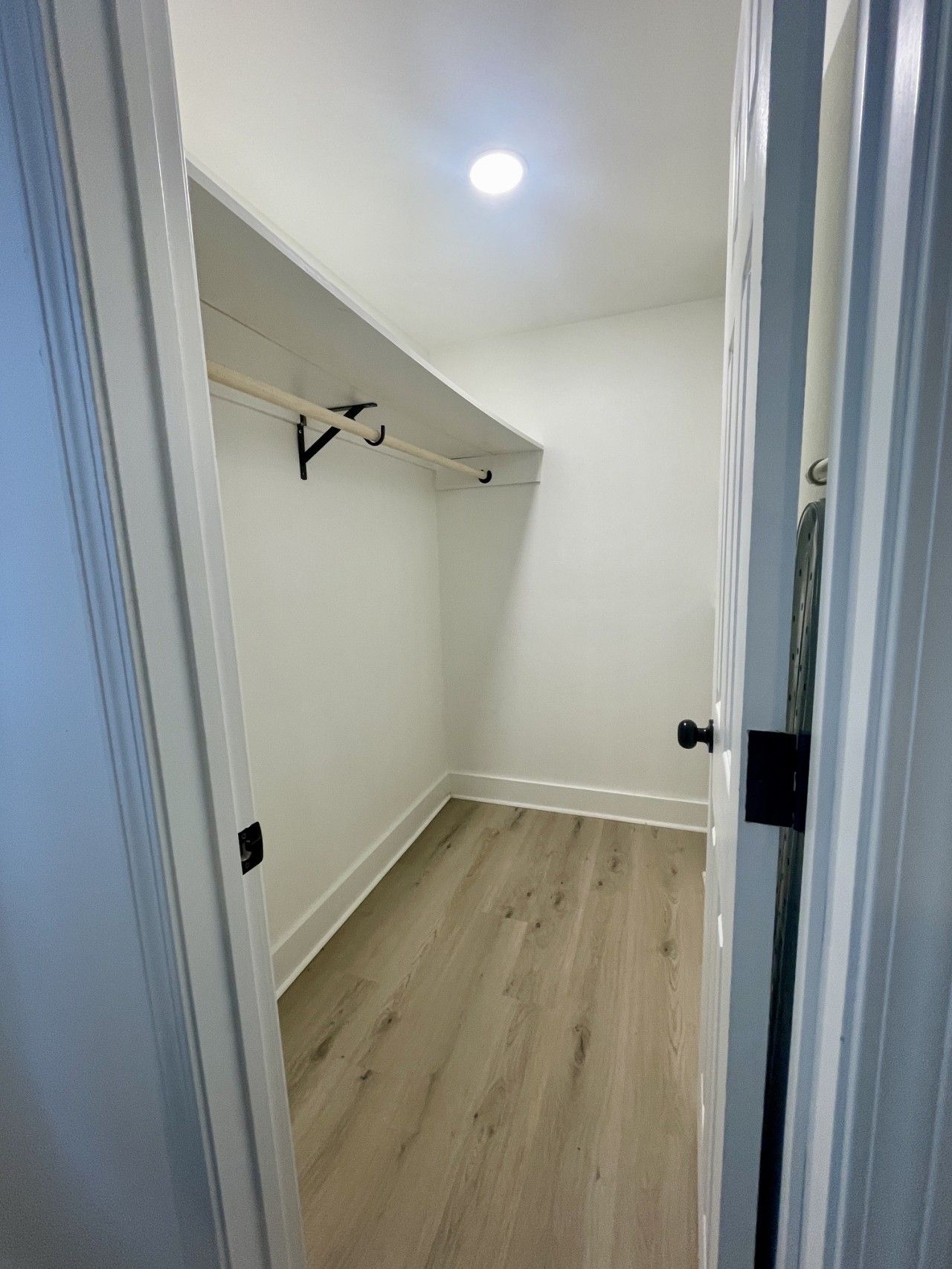 A walk-in closet with a wooden floor, white walls, and a ceiling light.