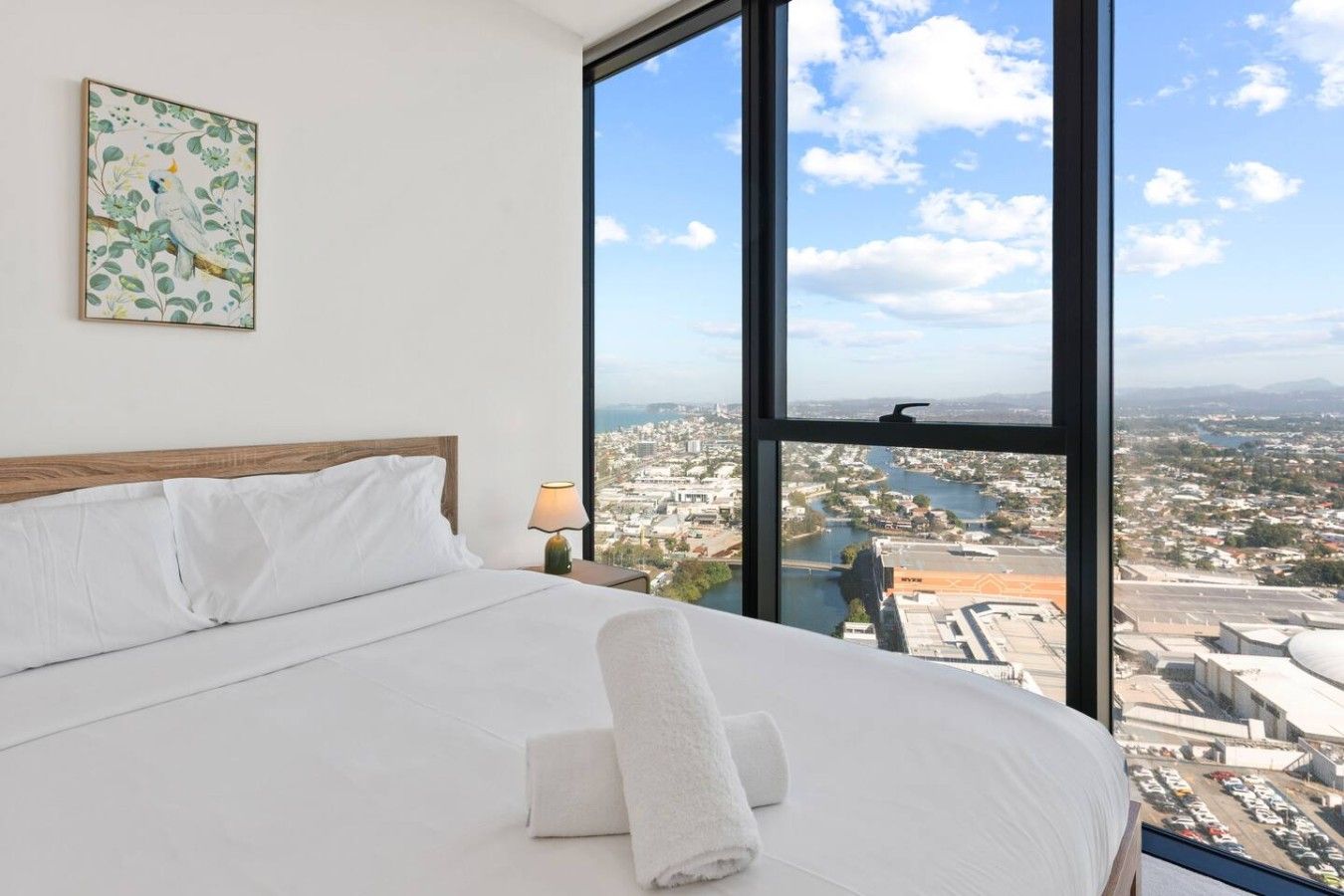 Habitación de hotel con un ventanal grande, cama con ropa blanca, lámpara y un cuadro en la pared.
