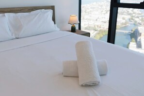 Ein ordentlich bezogenes Bett mit weißen Leinen und aufgerollten Handtüchern, eine Nachttischlampe und Blick auf eine Stadtlandschaft durch ein großes Fenster.