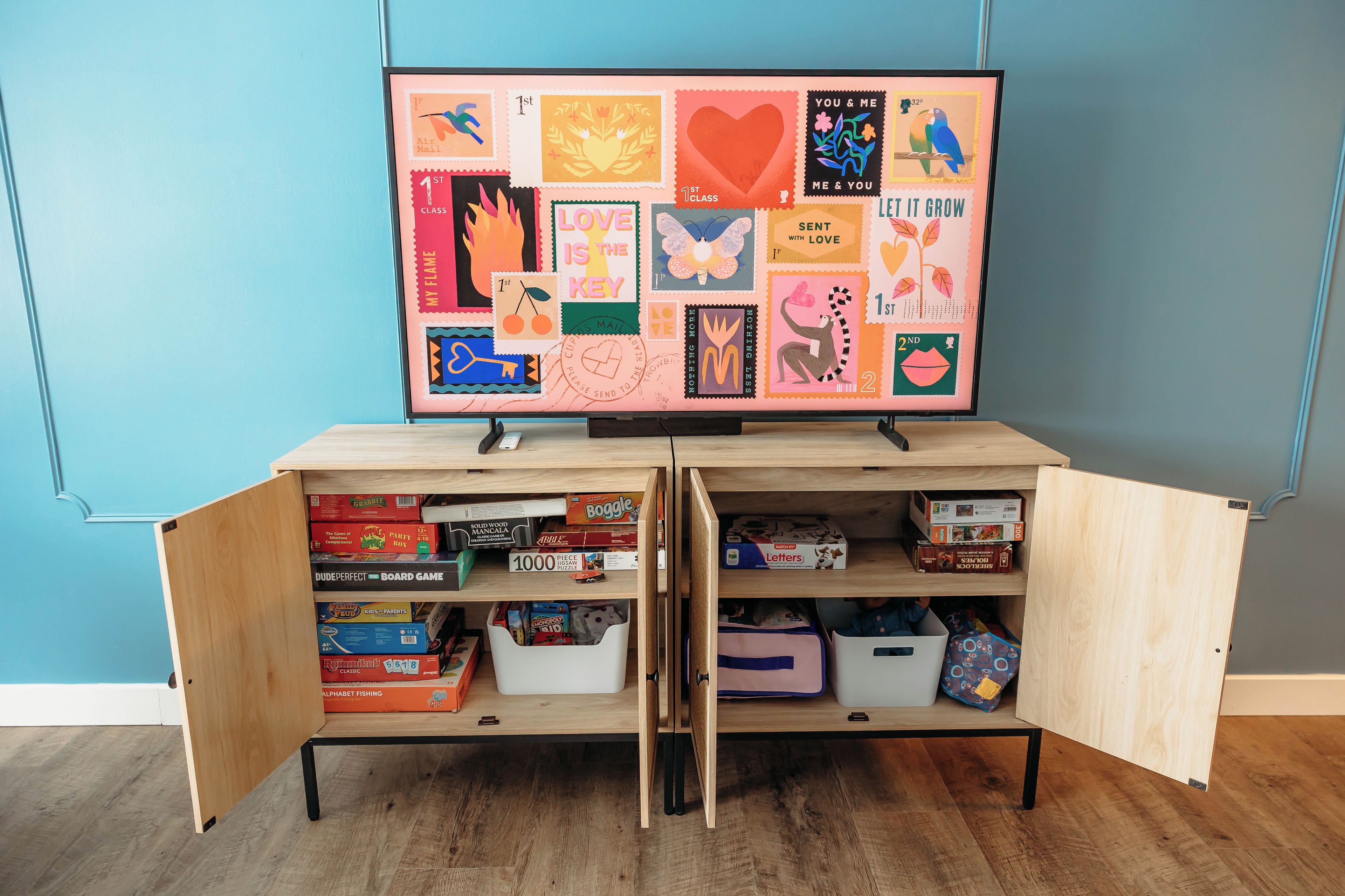 Ein Zimmer mit an der Wand montiertem Fernseher, der einen farbenfrohen Plakat zeigt. Darunter befindet sich ein Holzschrank mit Regalen, die verschiedene Brettspiele und Aufbewahrungsbehälter enthalten.
