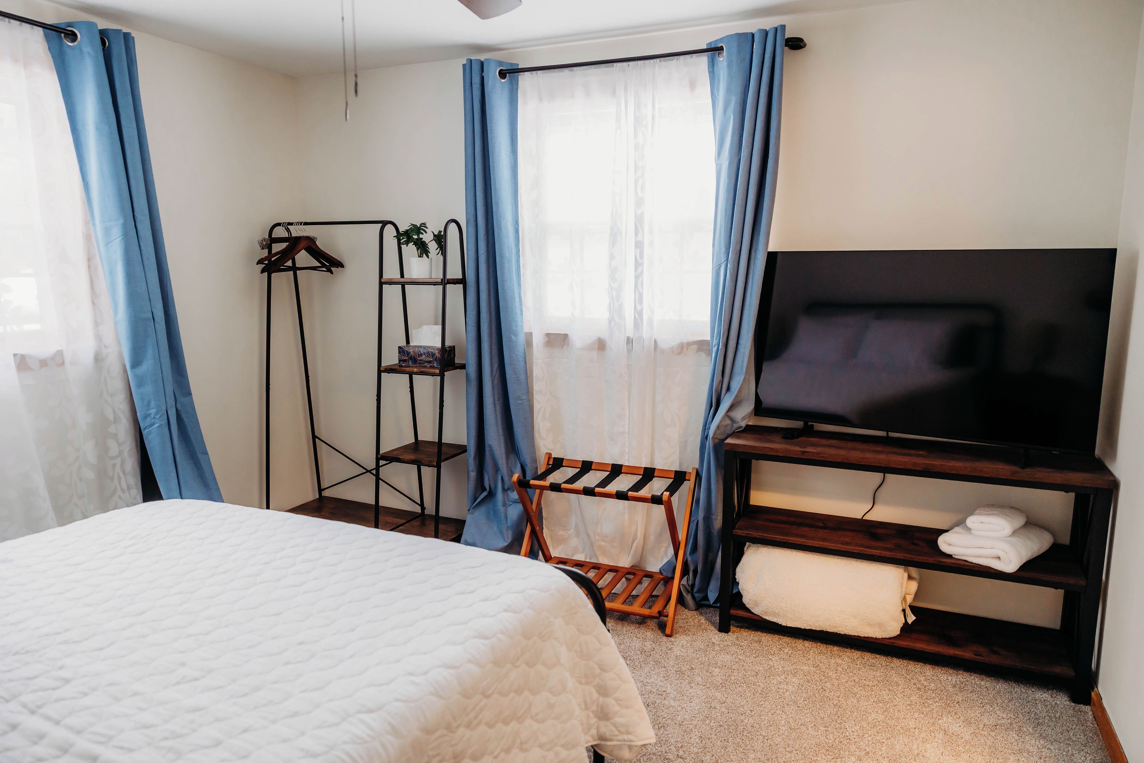 A bedroom with a bed, a TV on a wooden stand, a shelf with towels, and a plant on a metal rack.
