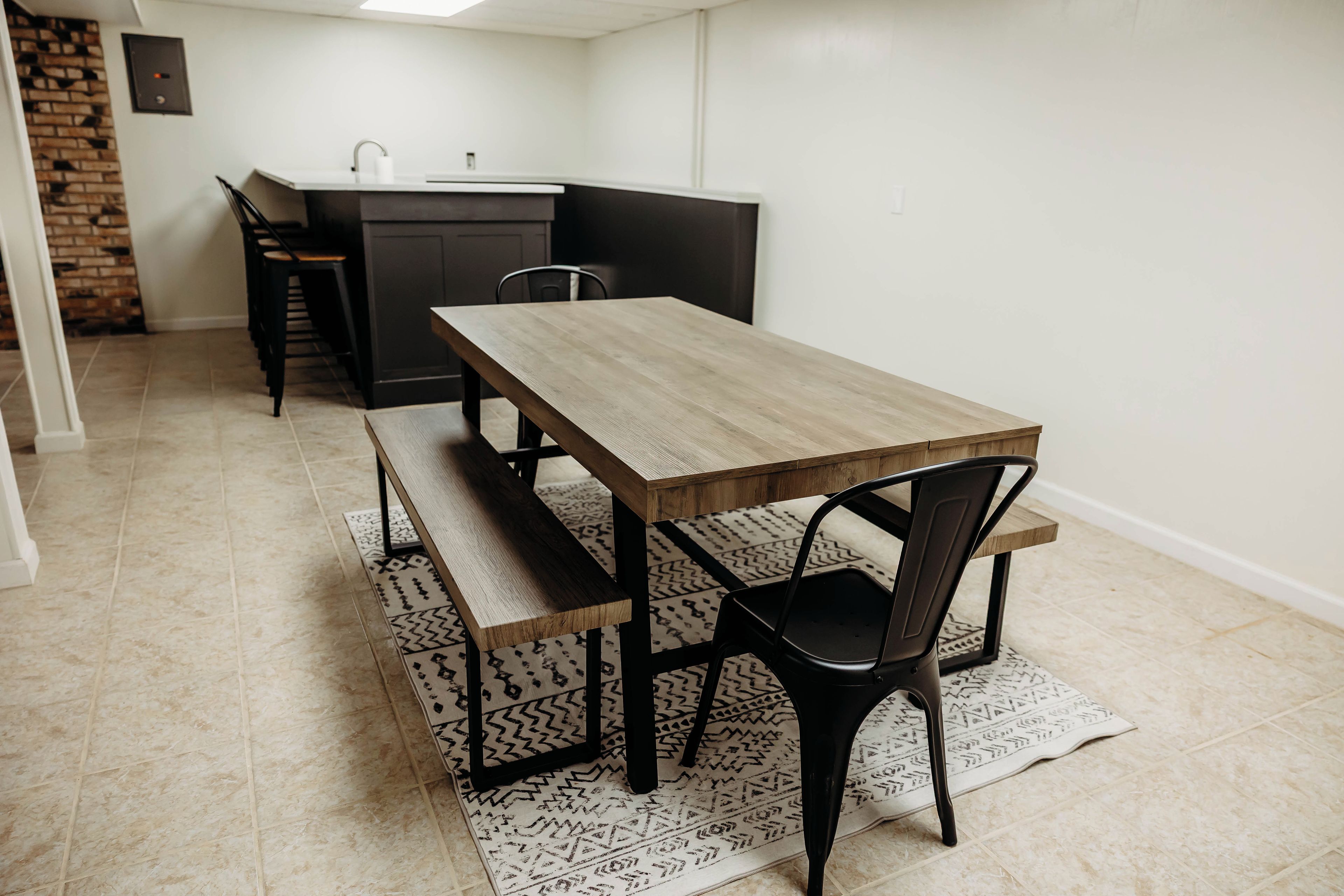 Zona pranzo con tavolo e panche in legno, cucina con lavello e tappeto decorato.