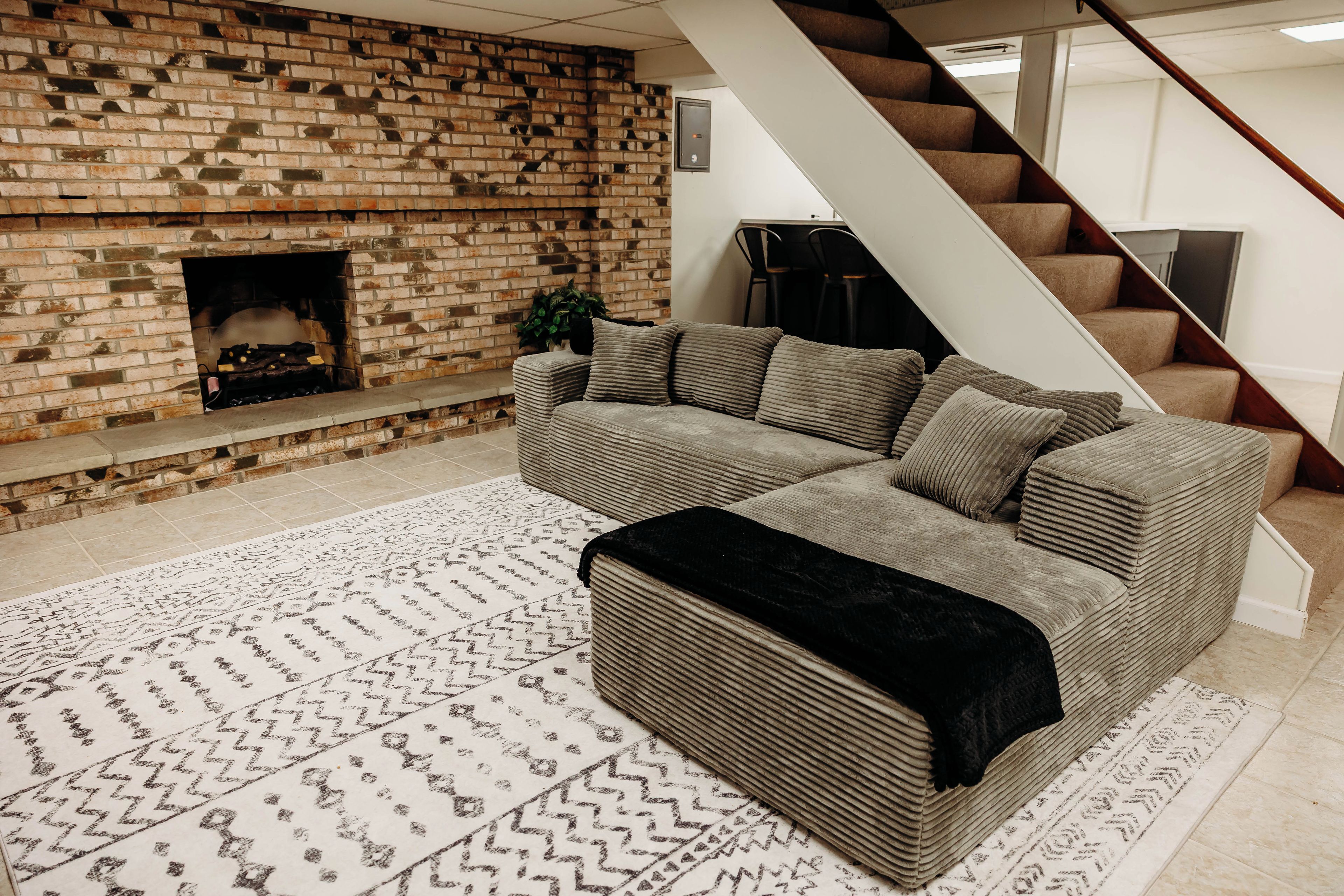 Una sala de estar con chimenea de ladrillo, un sofá modular, un otoman negro y una alfombra estampada.