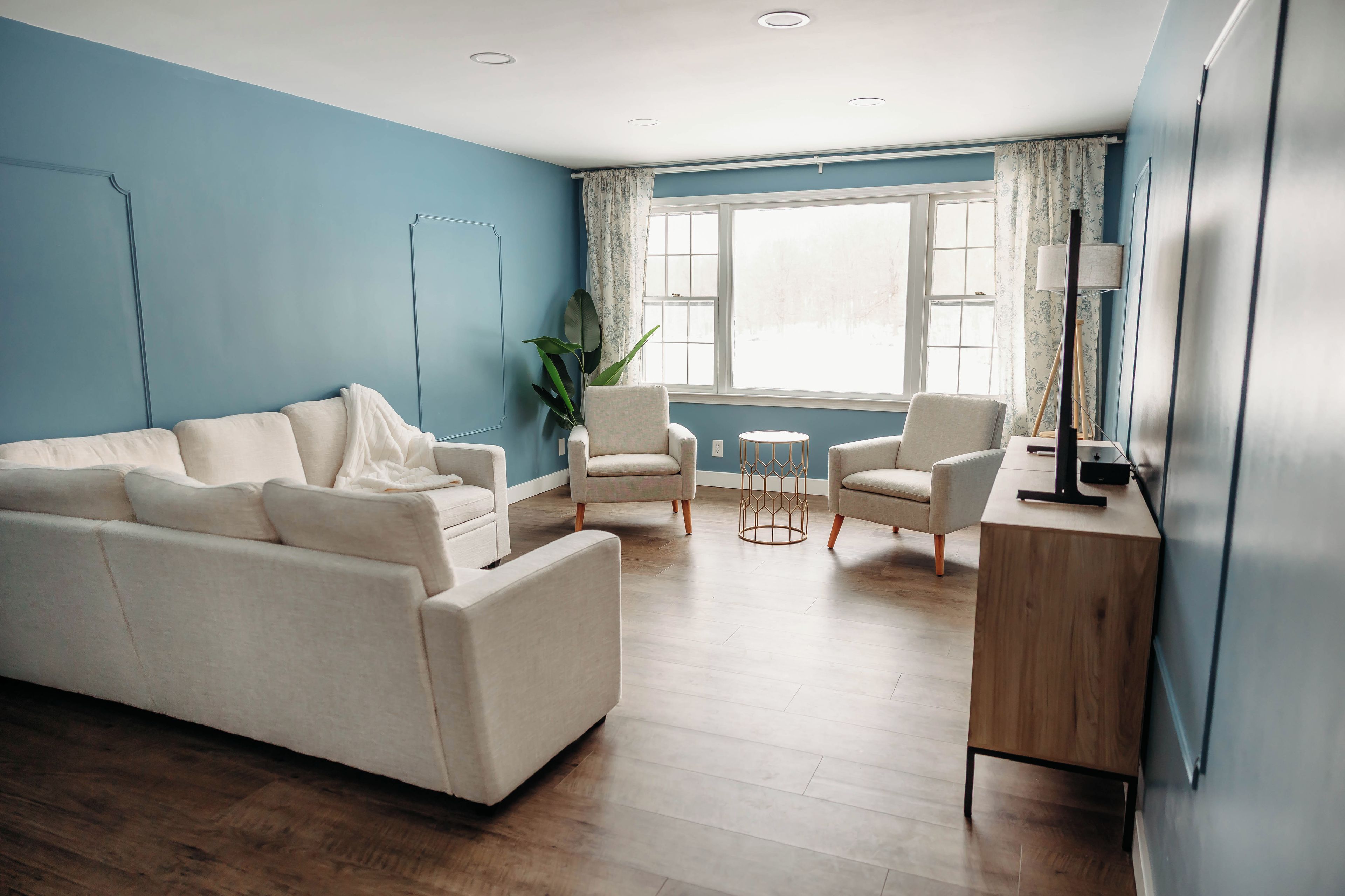 A modern living room with a blue accent wall, a beige sofa, two armchairs, a small round table, and a wooden desk.