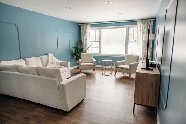 Una sala moderna con una pared azul de acento, un sofá color beige, dos sillones, una mesita redonda y un escritorio de madera.