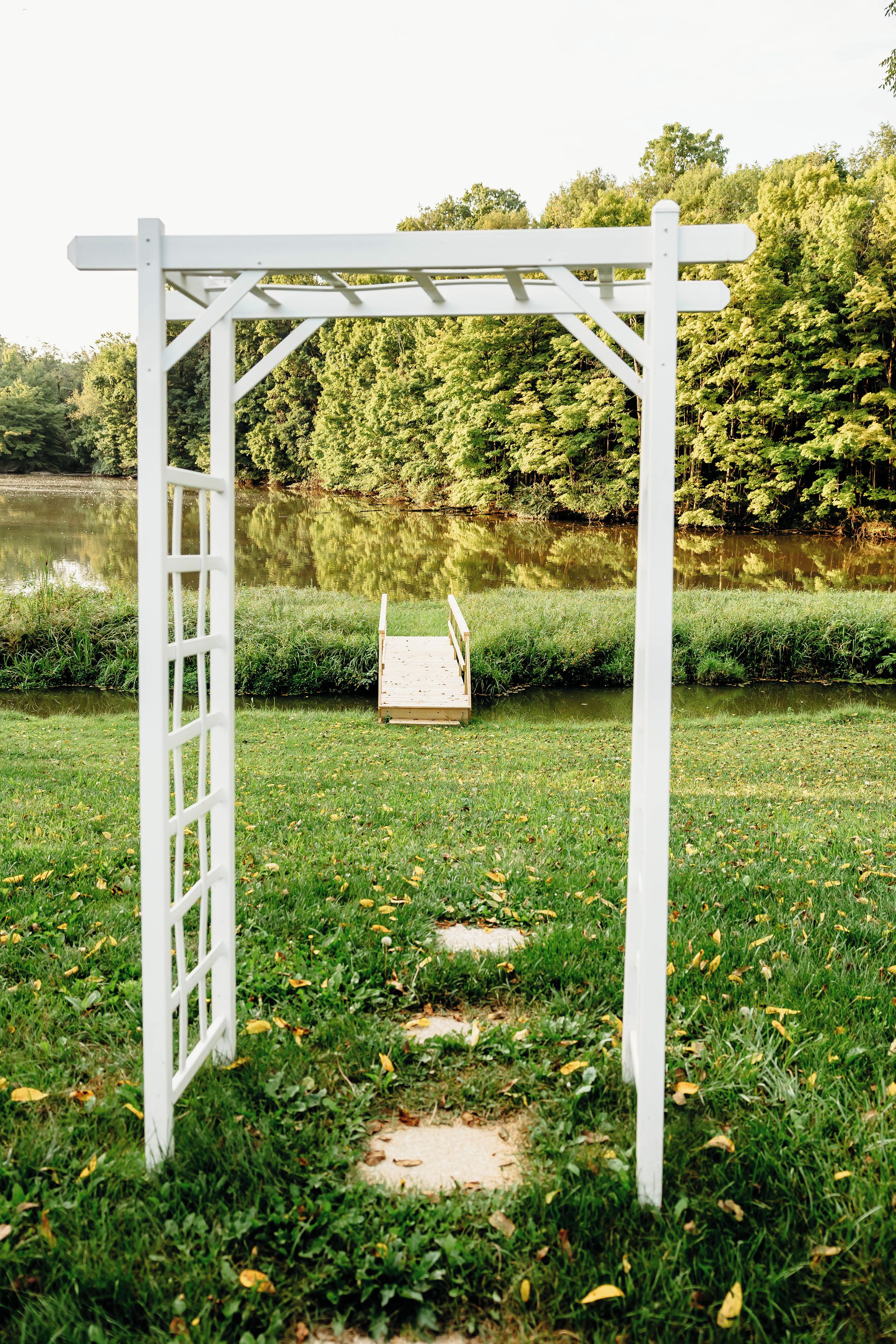 Una estructura de pérgola blanca con una plataforma de madera en un área cubierta de césped cerca de un cuerpo de agua.