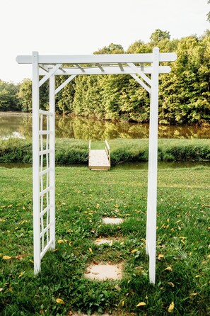 A white pergola structure with a wooden platform in a grassy area near a body of water.