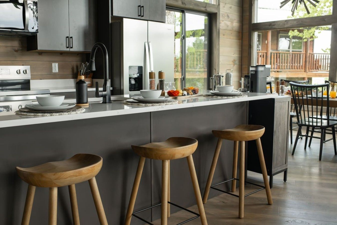A modern kitchen with a central island, bar stools, and a dining area visible through a large window.