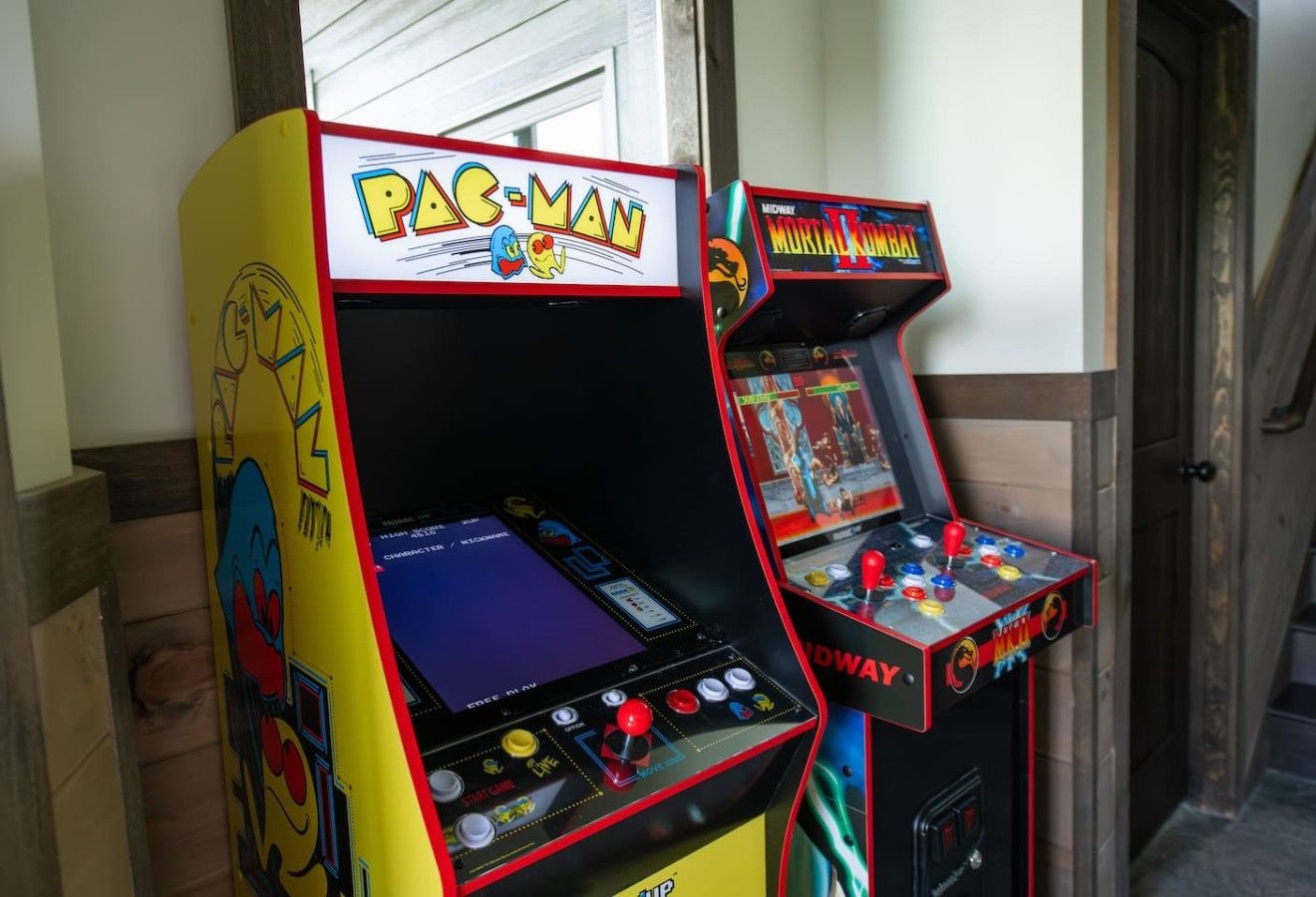 Two arcade machines, one labeled 'PAC-MAN' and the other 'Mortal Combat,' in a room with a wooden floor and a door in the background.