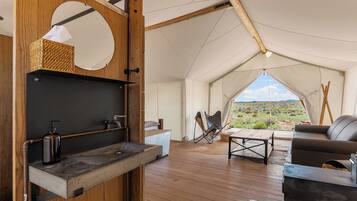 A tent interior with a wooden floor, a table, a sofa, and a view of the desert landscape.