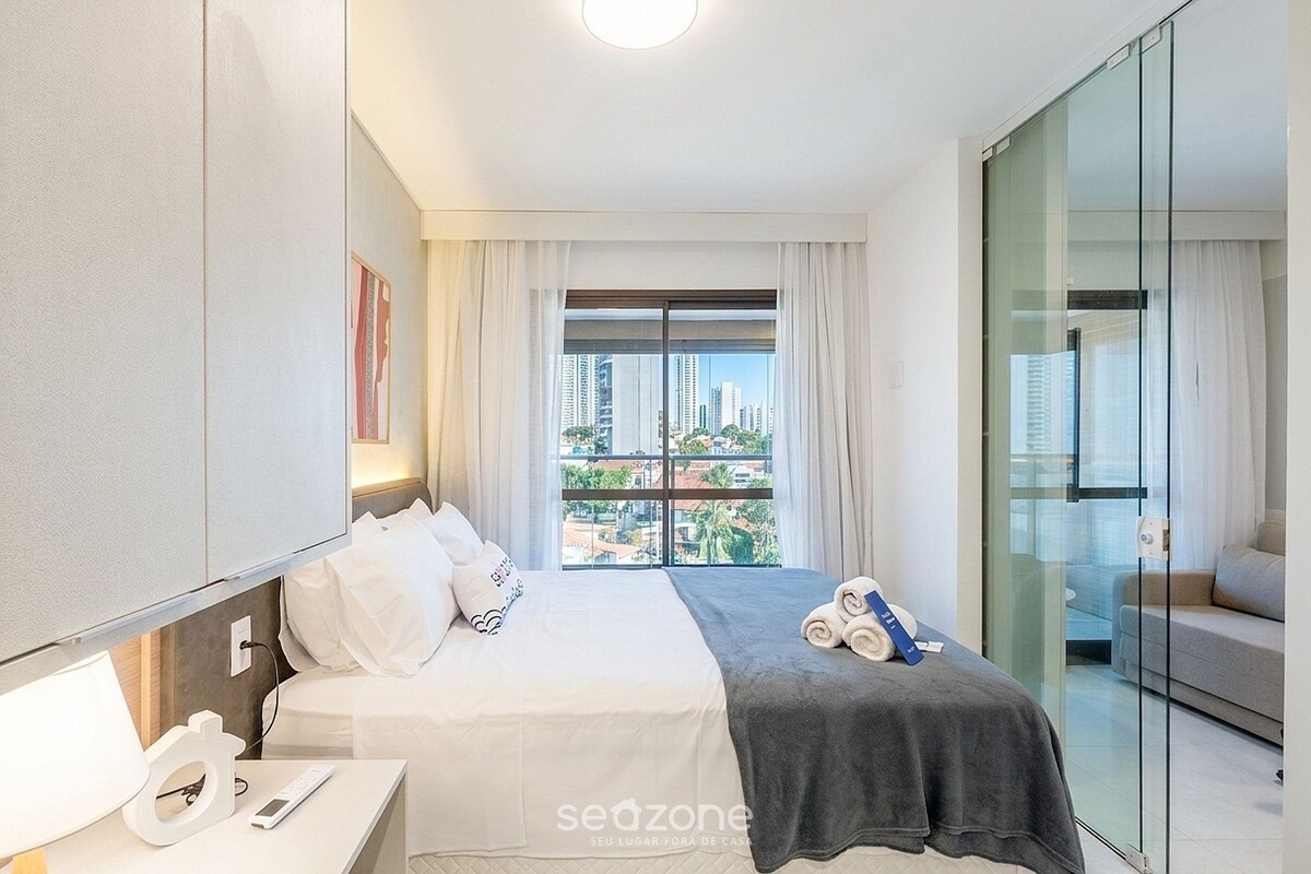 A modern hotel room with a bed, bedside table, and a view of the city through large windows.