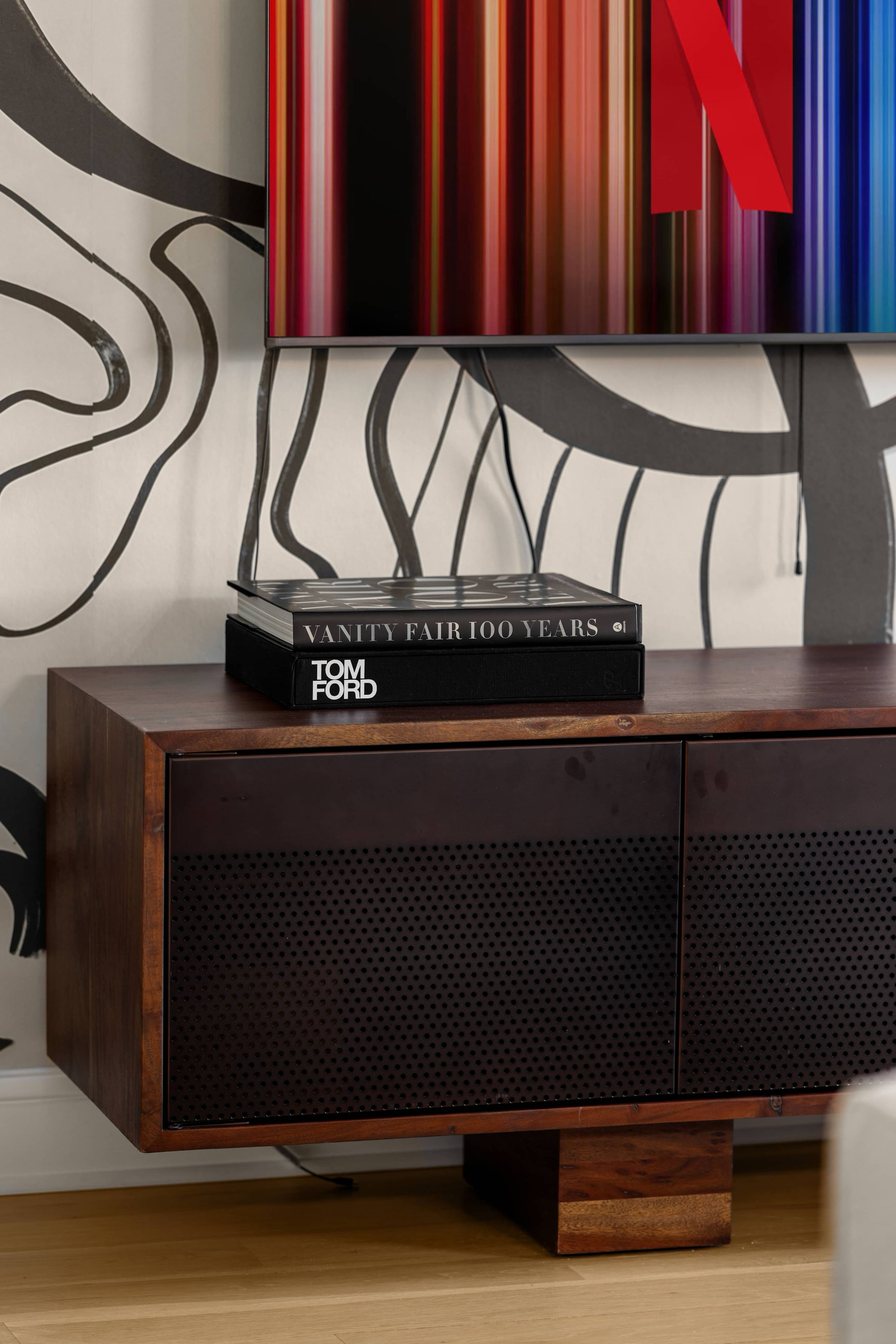 Een houten kast met een zwarte speakerrooster, een boek getiteld 'Vanity Fair 100 Years' van Tom Ford, en een kleurrijk abstract schilderij met een grote rode 'N' aan de muur.