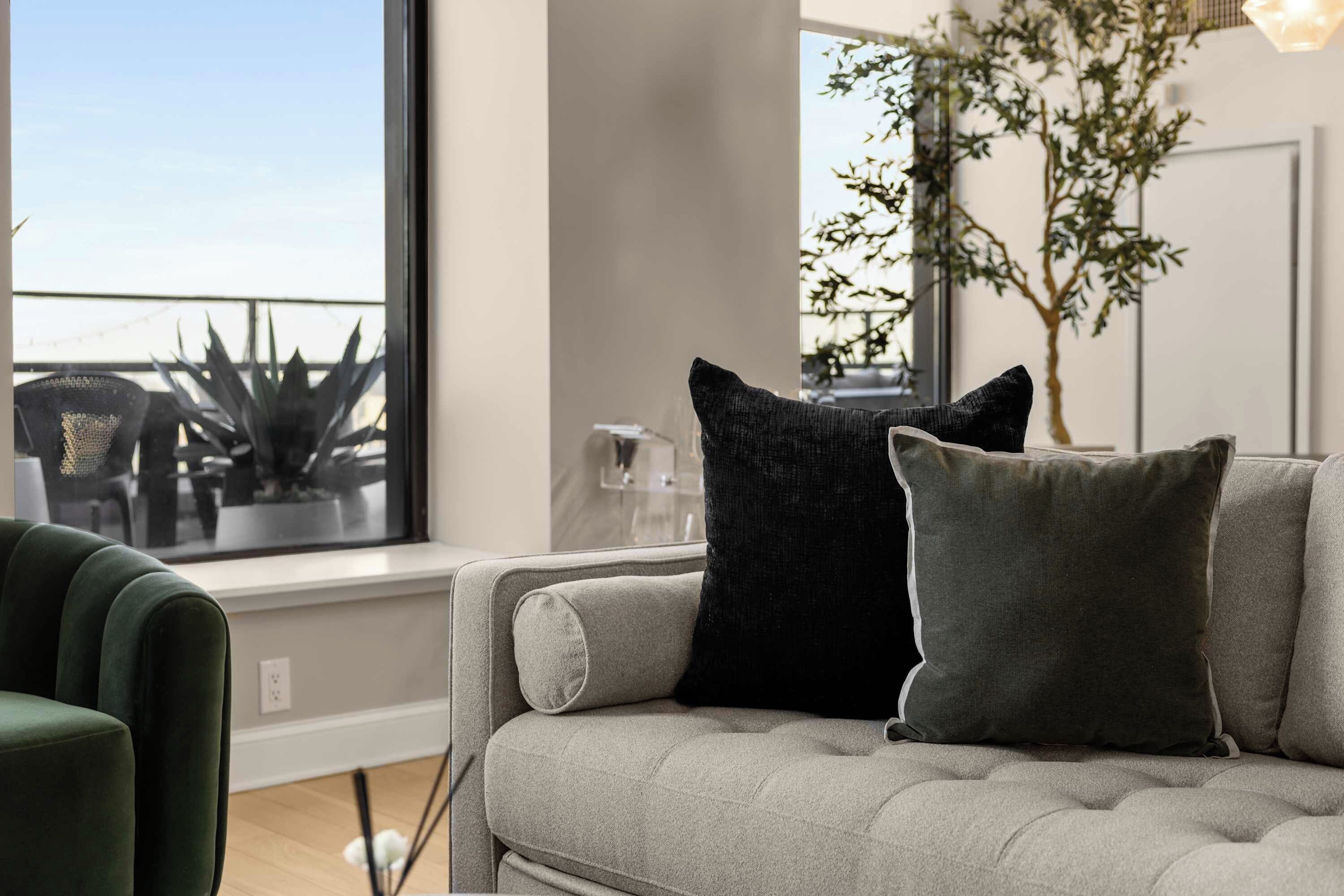 Een moderne woonkamer met een grijze bank, twee zwarte en één grijze kussen, een groene fauteuil, en een groot raam met uitzicht op een balkon en planten.