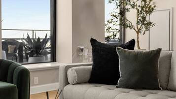 Een moderne woonkamer met een grijze bank, twee zwarte en één grijze kussen, een groene fauteuil, en een groot raam met uitzicht op een balkon en planten.