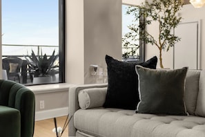 Een moderne woonkamer met een grijze bank, twee zwarte en één grijze kussen, een groene fauteuil, en een groot raam met uitzicht op een balkon en planten.