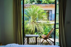 A room with a bed, a wooden table, and a chair. There is a window with curtains, and outside, there is a garden with various plants and a building. - Fresh Reno, 5 Min to Beach: Private Compound for Groups of 15 (Nosara)