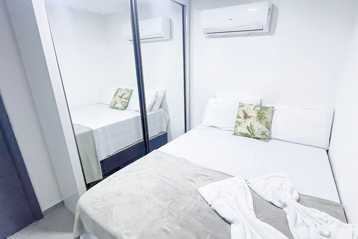 A hotel room with a bed, a bedside table, a mirror, and an air conditioning unit.