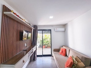 A modern living room with a flat-screen TV, a sofa with cushions, a sliding glass door leading to a balcony, and an air conditioning unit on the wall. - Residencial Di Mare - Porto de Galinhas (Ipojuca)