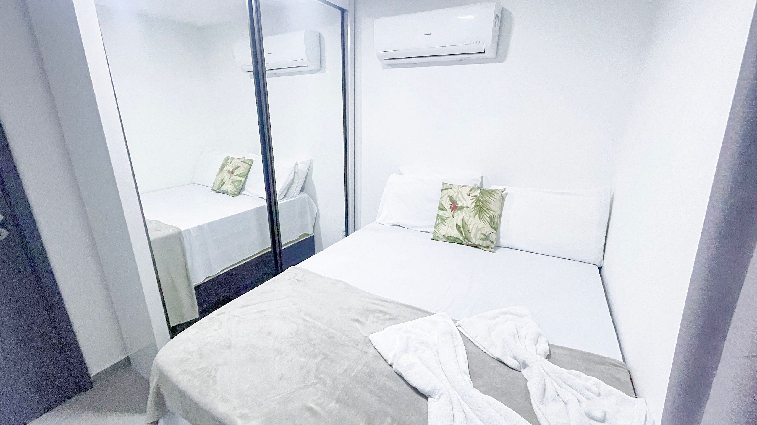 A hotel room with a bed, a bedside table, a mirror, and an air conditioning unit.