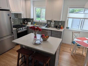 A modern kitchen with white cabinets, a stainless steel refrigerator, a gas stove, and a dining area with a red and white table. - Cozy 3 bed Craftsman Retreat, 10 Min from the Big House (Ann Arbor)