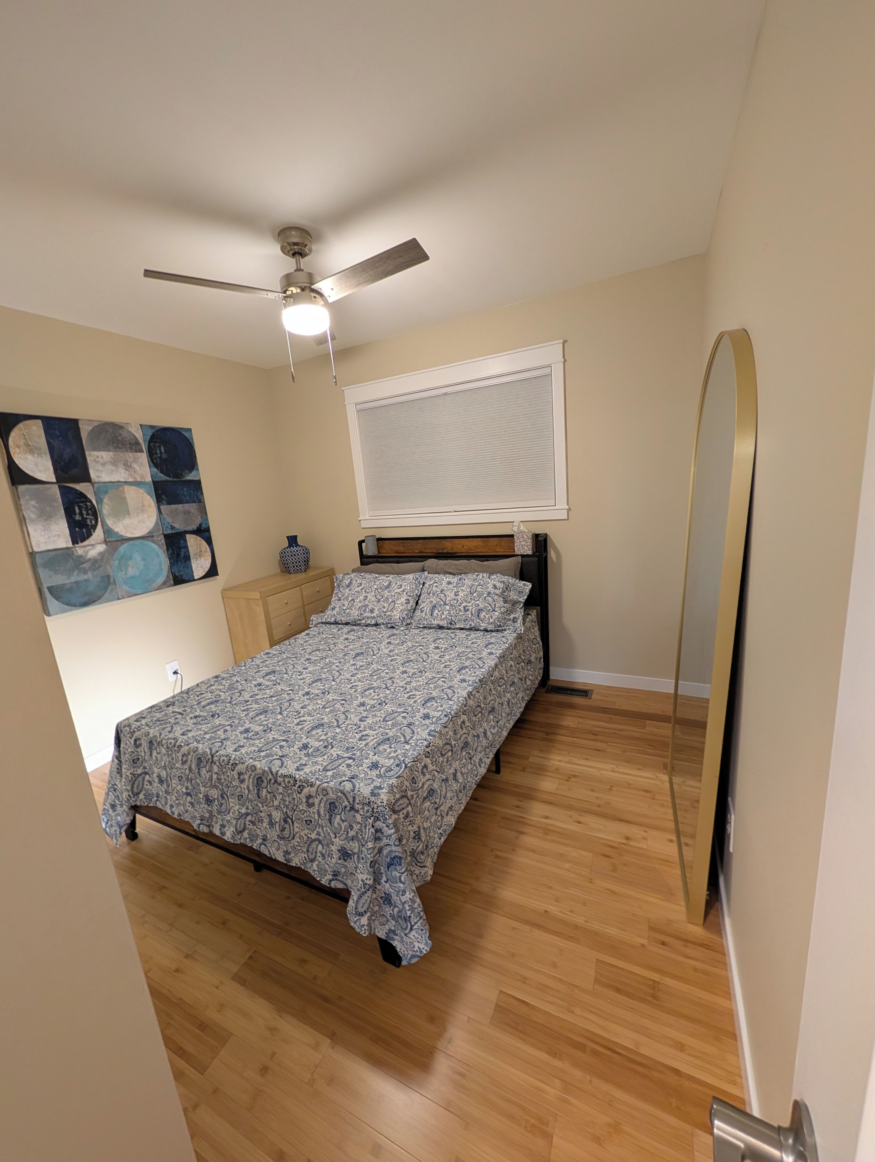A bedroom with a bed, a ceiling fan, a window with blinds, a wooden floor, and a wall-mounted mirror.
