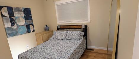 A bedroom with a bed, a ceiling fan, a window with blinds, a wooden floor, and a wall-mounted mirror.
