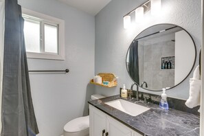 Een badkamer met een wit toilet, een wastafel met een marmeren blad, een ronde spiegel en een raam met gordijn.