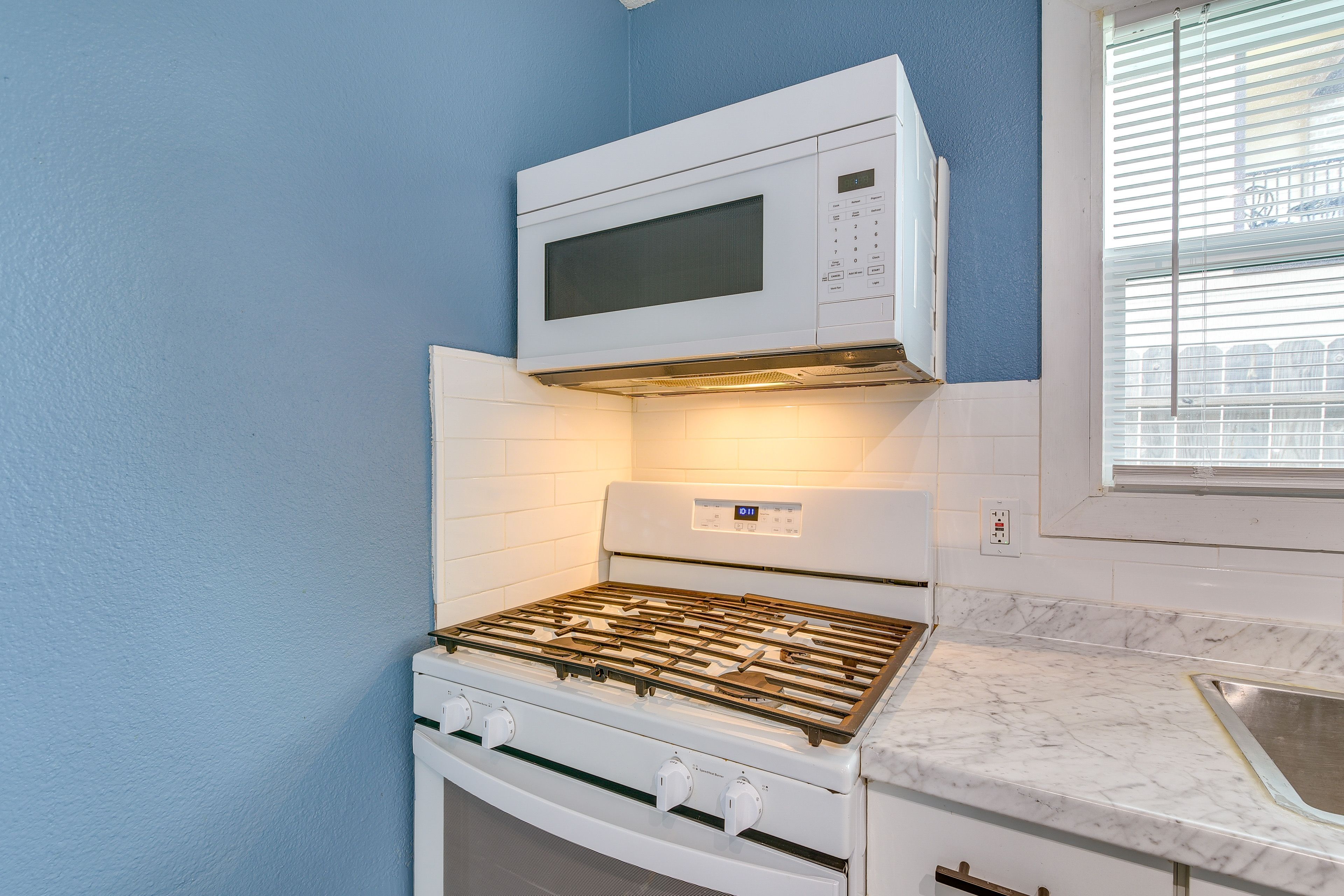 Cozinha com fogão, forno e micro-ondas brancos, janela com persianas e bancada de mármore.