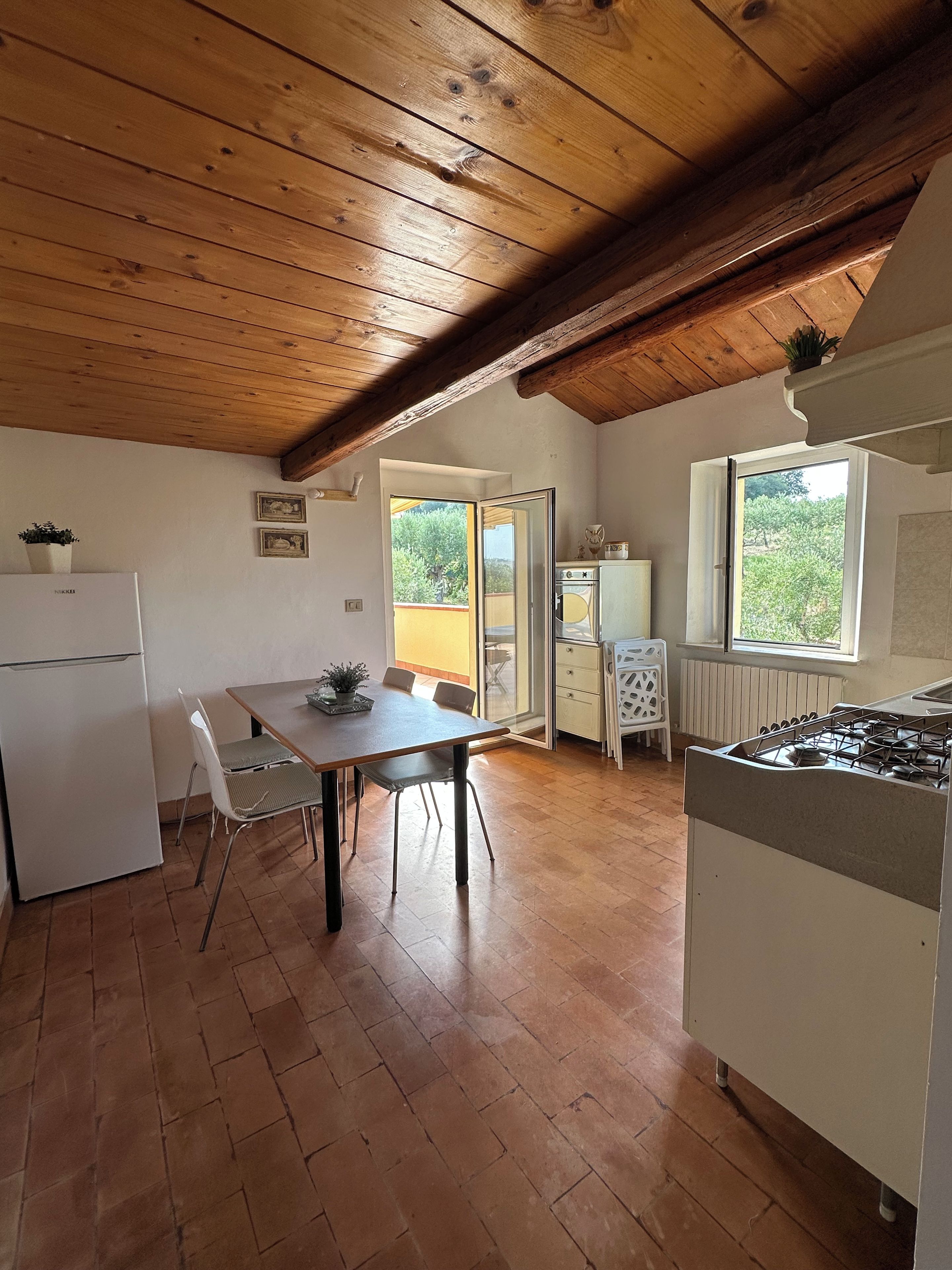 A kitchen with a wooden ceiling, a dining table with chairs, a refrigerator, and a window with a view of greenery.