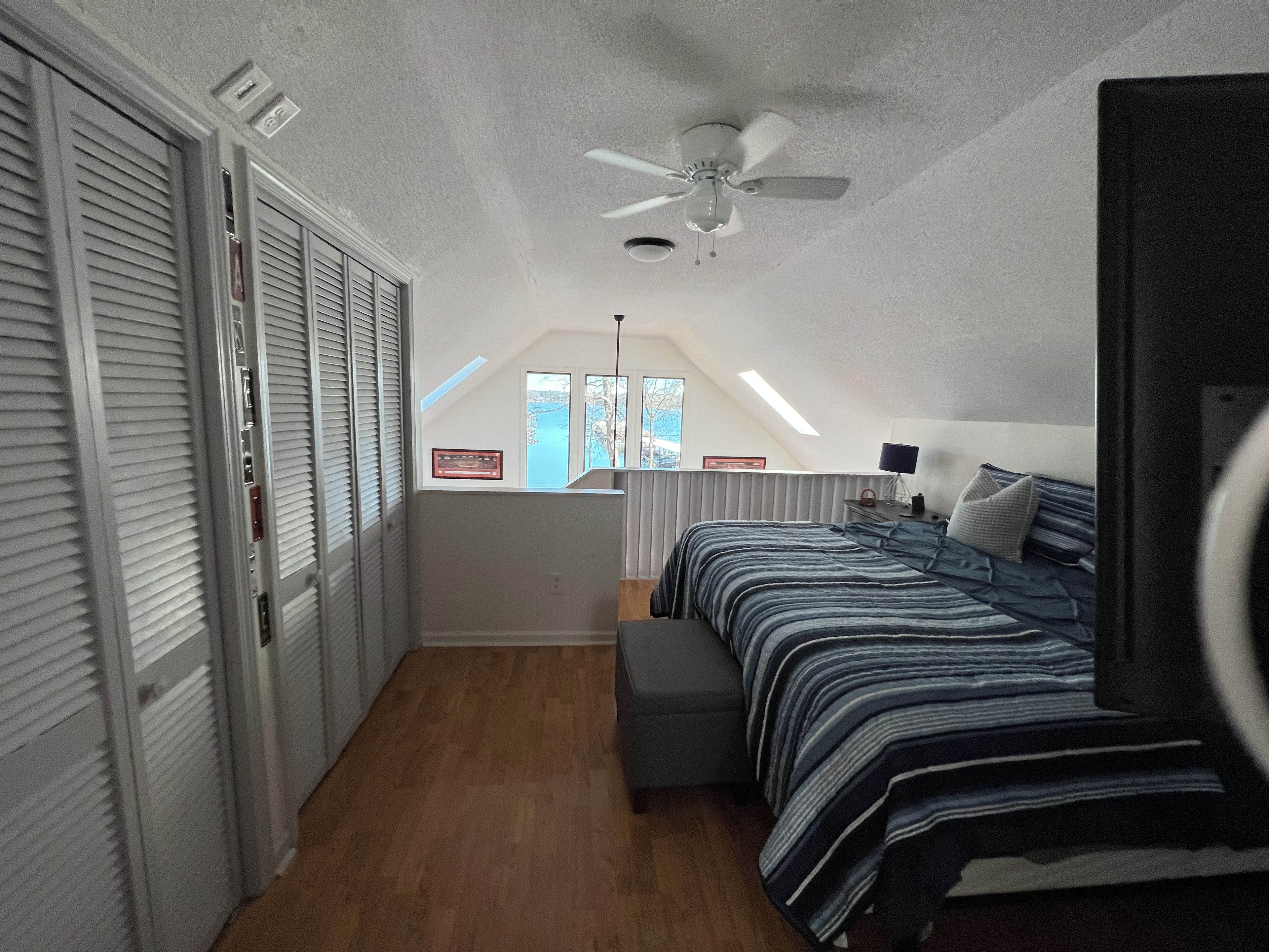 Une chambre avec un plafond en pente, un lit recouvert d’une couvre-lit à rayures, un ventilateur de plafond et une fenêtre donnant sur un beau paysage.