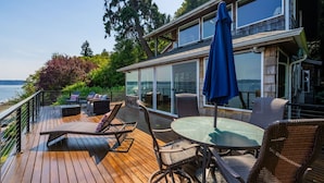 Une terrasse en bois avec des meubles d’extérieur, une pièce fermée par des baies vitrées et une vue sur l’eau.