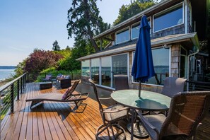 Eine Holzterrasse mit Gartenmöbeln, ein wintergartenartiger Raum und Blick auf das Wasser.